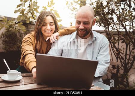 Cheerful couple of young people sitting at the cafe rooftop using laptop computer - new business concept with people working everywhere Stock Photo