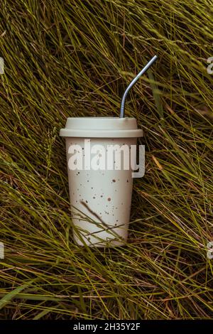 A beige reusable ceramic drink cup and a reusable straw lie on the green grass. Stock Photo