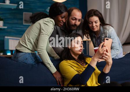 Multi-ethnic friends looking at fuuny videos on smartphone during movie night at home enjoying hanging out together. Group of people chilling in living room having fun. Friendship concept Stock Photo