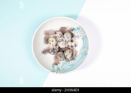 Easter table setting concept with quail eggs and feathers on plate decorated spring flowers on pastel white and light blue backdrop. Passover minimal Stock Photo