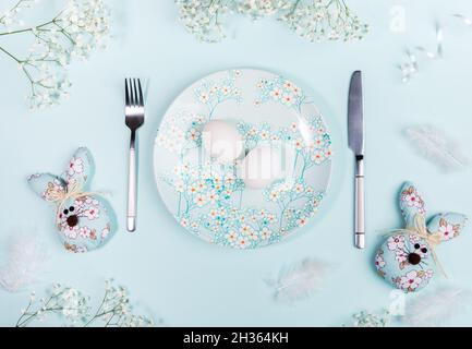 Flat lay Easter table setting with white eggs on plate with spring flowers decor, DIY textile Easter bunnies, feathers and white flowers on pastel blu Stock Photo