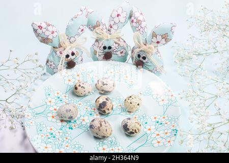 Easter composition. Three DIY textile Easter bunnies standing around plate with spring floral decor with quail eggs on pastel light blue. Horizontal o Stock Photo