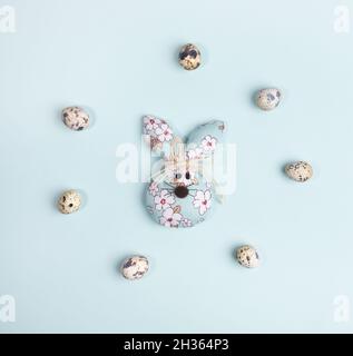 Flat lay Easter symbols arrangement. Quail eggs laid out in circle around one DIY textile Easter bunny on pastel light blue. Festive layout. Square ph Stock Photo