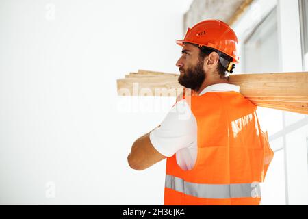 Builder man in hardhat carrying timber on building site, close up . Stock Photo