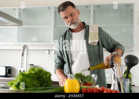 Mature man cooking vegetables salad in home kitchen. Male hands pouring olive oil in bowl with salad. Stock Photo