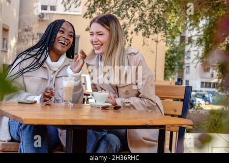 Two young girlfriends enjoying coffee together on a terrace of the coffee shop, sitting at a table, chatting and laughing out loud. Diversity. Stock Photo