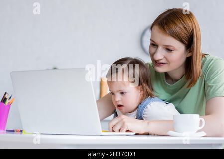 Smiling freelancer using laptop near daughter with down syndrome and coffee at home Stock Photo