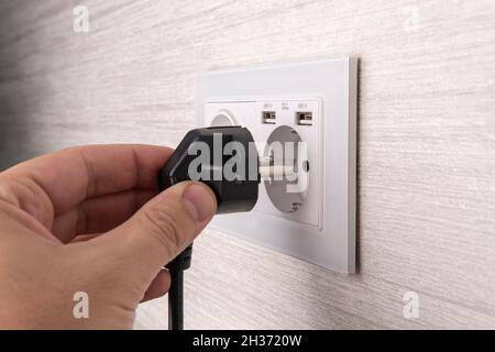 Hand plugging in an electric cord into a white plasticmodern socket or european wall outlet on grey wall. Closeup of a man hand inserting an electrica Stock Photo