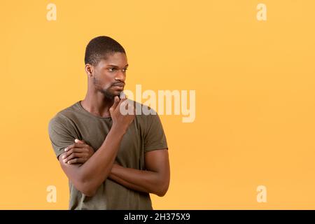 Pensive african american man touching chin and looking aside at free space while standing over yellow background Stock Photo