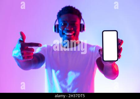 Check out new music app. Happy black guy in headphones pointing at smartphone with empty screen in neon light, mockup Stock Photo