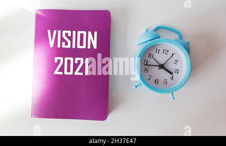 Text Vision 2021 is written on a crimson notepad that lies on a white background next to an alarm clock Stock Photo