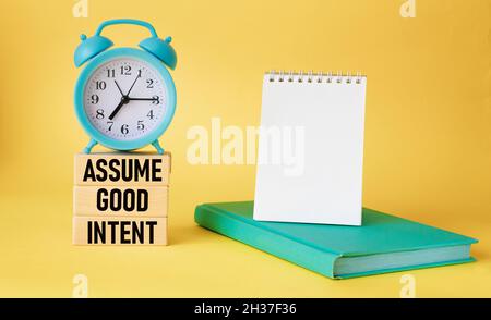 Assume good intentions. Inspirational quote on wooden blocks with notepad to write on yellow background with alarm clock. Positivity concept Stock Photo