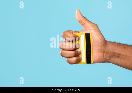 Closeup view of African American man holding credit card and showing thumb up on blue studio background, copy space Stock Photo