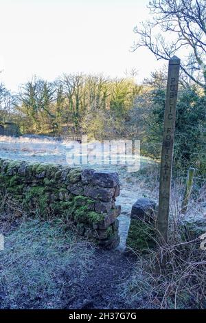 A wooden footpath marker and stone stile with the path leading to a wooden Bridge through a field covered in frost, Winter, Staffordshire, England, UK Stock Photo