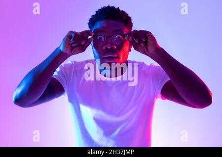 Young black guy touching his glasses, opening his eyes wide in shock in neon light Stock Photo
