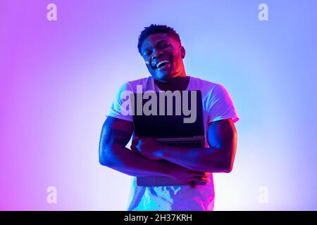 Technology lover concept. Happy black man hugging laptop with blank screen in neon light, mockup for website design Stock Photo