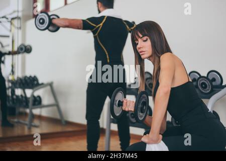 Determined young couple working out in gym. Fit boy and girl doing exercise with weights. Beautiful sporty woman. Strong boys lifting the dumbells in Stock Photo