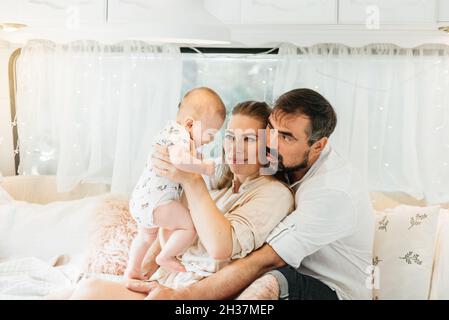 Family with baby on the bed in trailer in the morning. Newborn child traveling in camper with parents. View from van inside Stock Photo