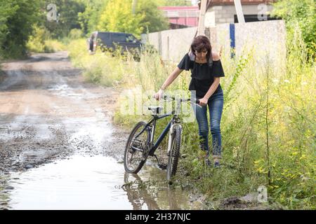 A woman with difficulty drags a bicycle through the mud, afraid to drive through a puddle. Stock Photo