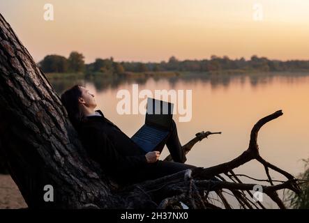 Handsome businessman in stylish black suit sits on tree trunk by the water with laptop in his hand at sunset.  Stock Photo