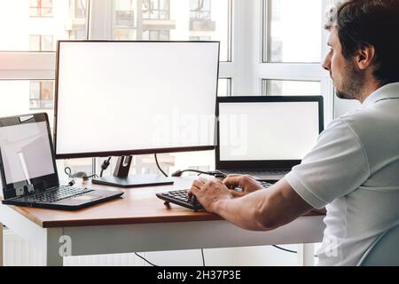 Man sitting at workplace with two laptops and monitor near the window. Remote work from home Stock Photo
