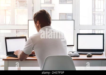 Man sitting at workplace with two laptops and monitor near the window. Remote work from home Stock Photo