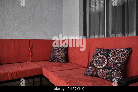 Vintage style sofa with red color sitting area and colorful patterned pillows on it with big windows and plastered wall background. Stock Photo