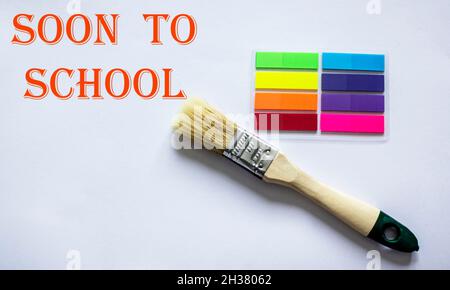 Written on the blackboard soon to school, surrounded by colored stickers and a paint brush on a white background Stock Photo
