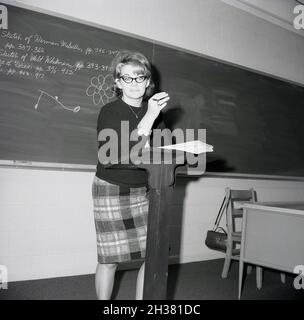 1960s, historical, a lady teacher standing with her notes at a wooden lectern in a classrooom infront on a blackboard, Virginia, USA. Written in chalk on the blackboard the names of two famous American writers, Walt Whitman and Herman Meville. Stock Photo