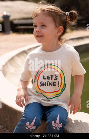 little beautiful red-haired girl in a T-shirt laughs while sitting on the bank of the fountain Stock Photo