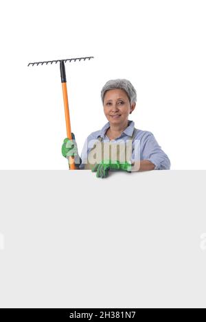Mature asian woman gardener with rake standing behind blank banner with empty copy space for text isolated on white background Stock Photo
