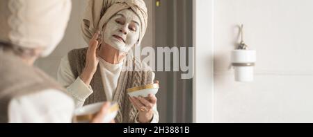 lderly woman with towel on her head smiles and applies cleansing mask . Stock Photo