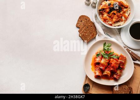 Bowl with delicious oatmeal, beef stew and plate with tasty prunes baked in crispy bacon, gravy and parsley on light background Stock Photo