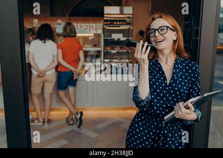 Redhead young woman makes voice call keeps smartphone near mouth wears spectacles polka dot dress Stock Photo