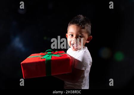 Happy boy with Christmas present in his hands, on black background. Beautiful latin boy isolated with red present. Stock Photo