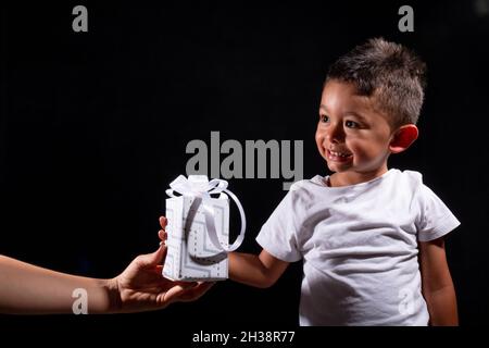 Child receiving gift from his mother, black background. Woman hand handing white gift to small child and isolated on black background. Stock Photo