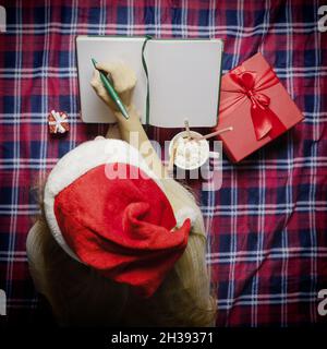 A girl in a Santa hat makes notes in a diary while lying on a plaid blanket surrounded by Christmas gizmos. Top view Stock Photo