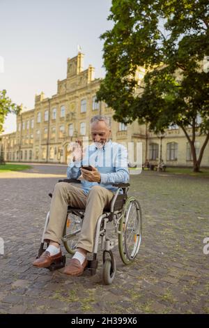 Smiling senior man, recovering male patient in wheelchair making a video call using smartphone while resting alone in the park near hospital Stock Photo