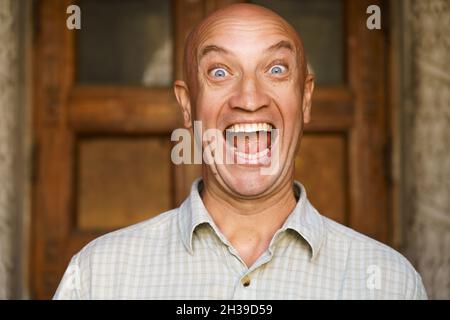 Portrait of an emotional bald guy with blue eyes. Dressed in light shirt. Shows different grimaces close-up on the background of the old door. Caucasian man with facial expressions Stock Photo