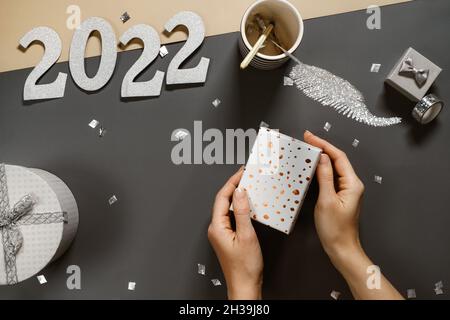 Hands wrapping box on dark grey desk with small gift, confetti. Getting ready for New Year celebration. Minimalistic stylish silver table top view Stock Photo