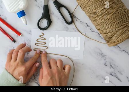 DIY ribbon rope, scissors used to create individual greeting card merry christmas. Christmas tree made of rope. Top view of hands making step by step Stock Photo