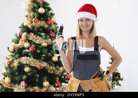 Woman builder in red santa claus hat holding drill in hands near christmas tree Stock Photo