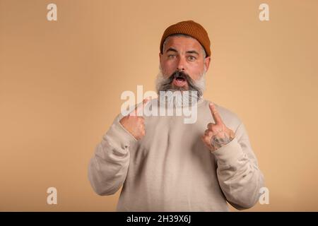 Hipster man looking disappointedly at camera pointing at himself with hands being surprised to be choosed by someone. Stock Photo