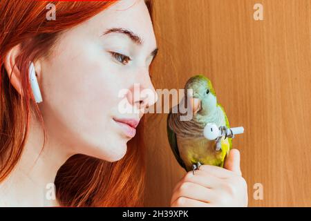 A girl with red hair and a earphone in her ear is holding a green parrot. Stock Photo