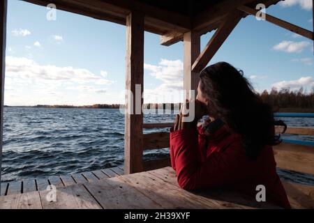A woman in a Red Jacket Sits at a Table on a Wooden Pier and Looks at the Water, Clouds and Trees with Yellow Foliage. Autumn Landscape. Stock Photo