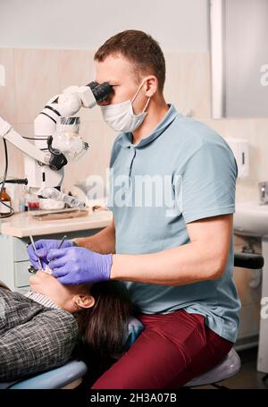 Dentist in medical mask using dental microscope while treating patient teeth. Woman lying in dental chair while having dental check-up in modern medical center. Concept of dentistry and dental care. Stock Photo