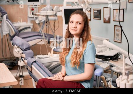 Portrait of charming female dentist with long red hair posing in dental office with dental chair and special equipment. Concept of dentistry, healthcare and dental staff. Stock Photo