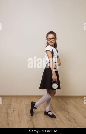 Full length portrait of young school girl holding alarm clock and thinking over light background. School timing concept. Knowledge day. Stock Photo