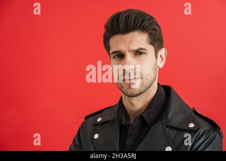 Young unshaven man wearing leather jacket looking at camera isolated over red wall Stock Photo