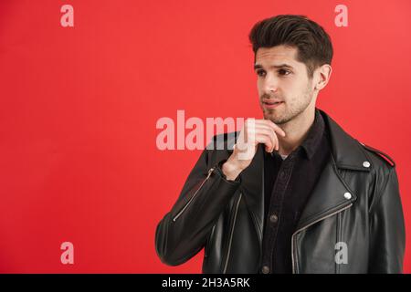 Young unshaven man wearing leather jacket looking aside isolated over red wall Stock Photo